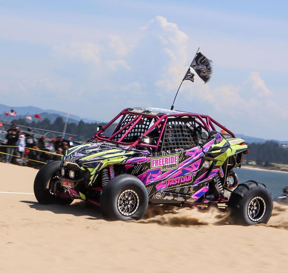 Hostile H333 Glamis Beadlock wheels on a purple and green Polaris RZR Pro R kicking up sand at a dune event. Hostile gallery.